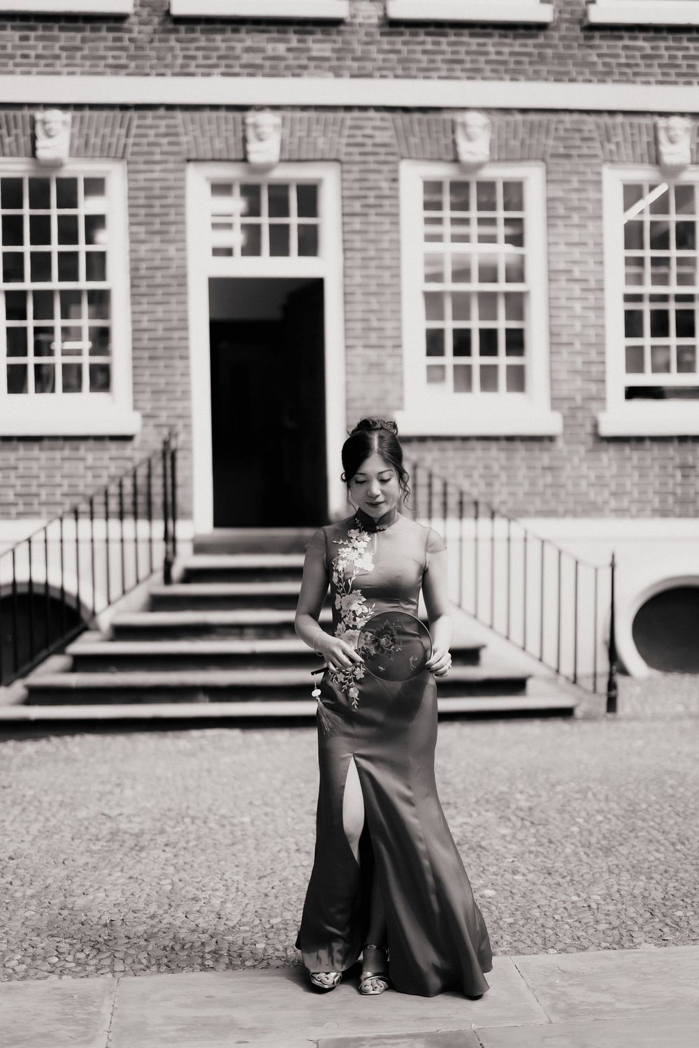 Woman in elegant floor-length gown stands before a classic brick building in this black and white portrait.