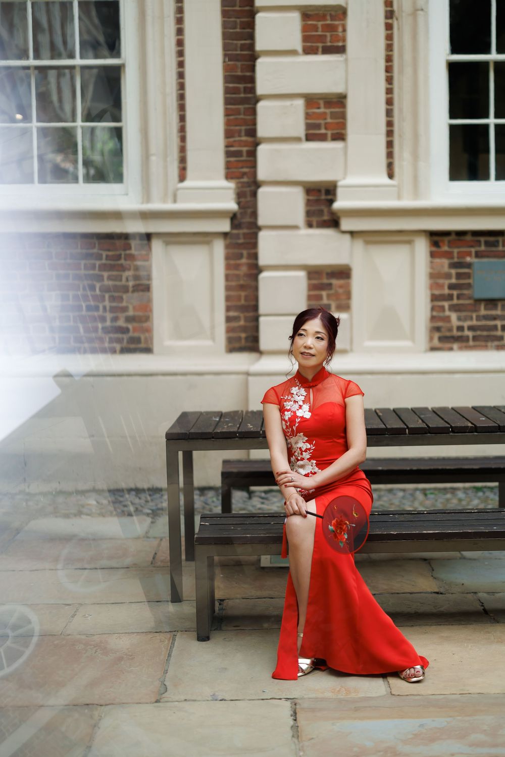 Woman in red embroidered cheongsam sits on bench outside brick building, holding decorative fan.