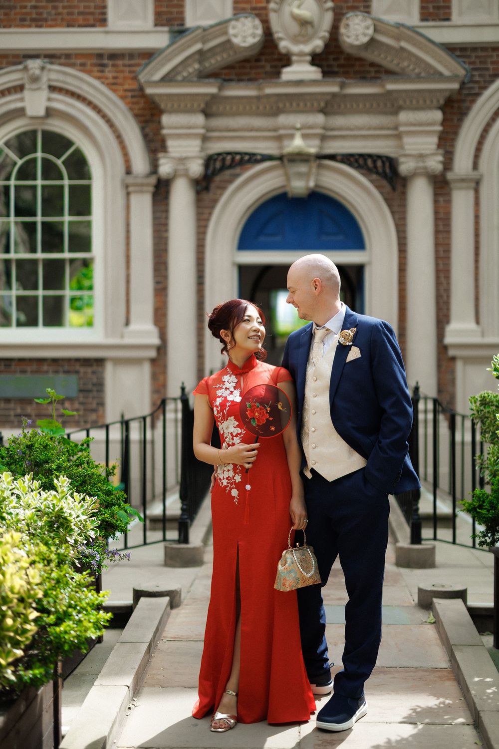 Couple in red qipao and navy suit posing on steps outside elegant brick building on wedding day.