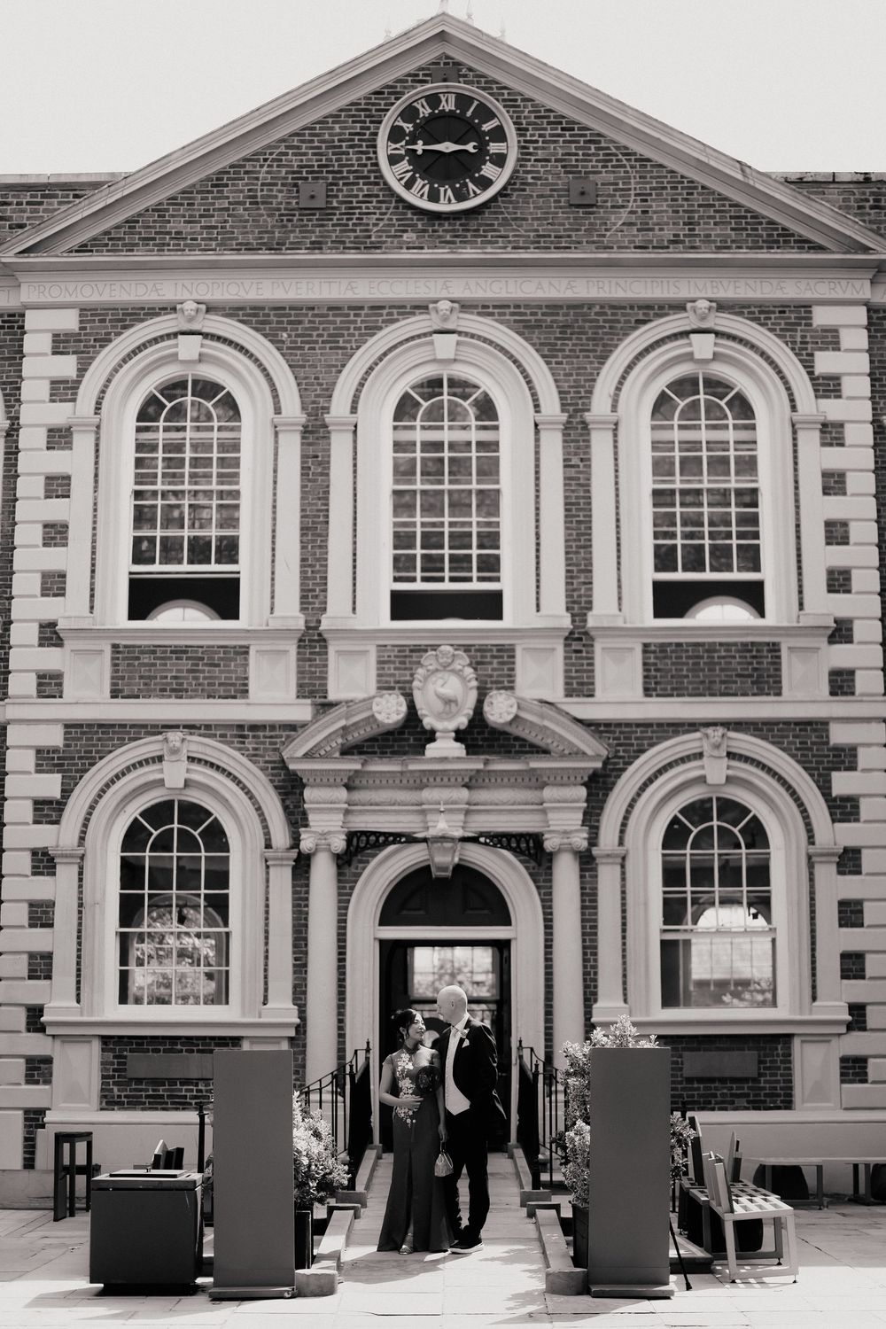 A bride and groom pose outside a grand Georgian brick building with arched windows and a clock tower.
