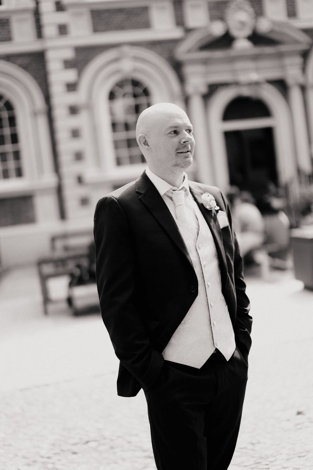 Bald groom in black tuxedo and ivory waistcoat stands confidently outside elegant venue in black and white photo.