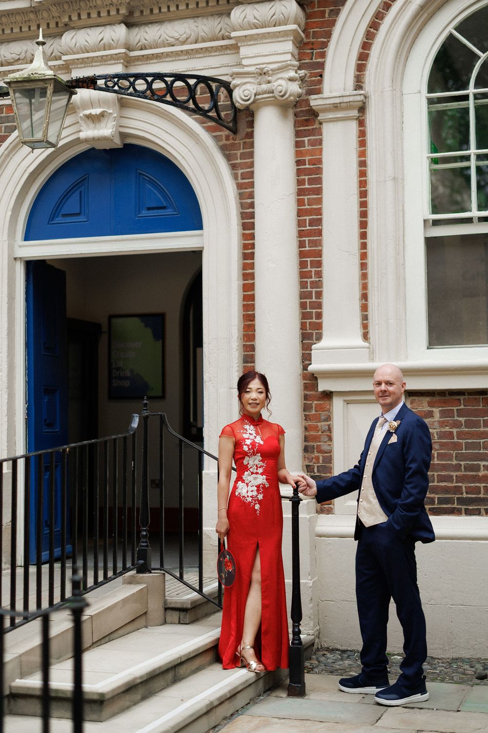 Couple posing outside brick building, woman in red Chinese qipao dress, man in navy suit, by blue arched door.