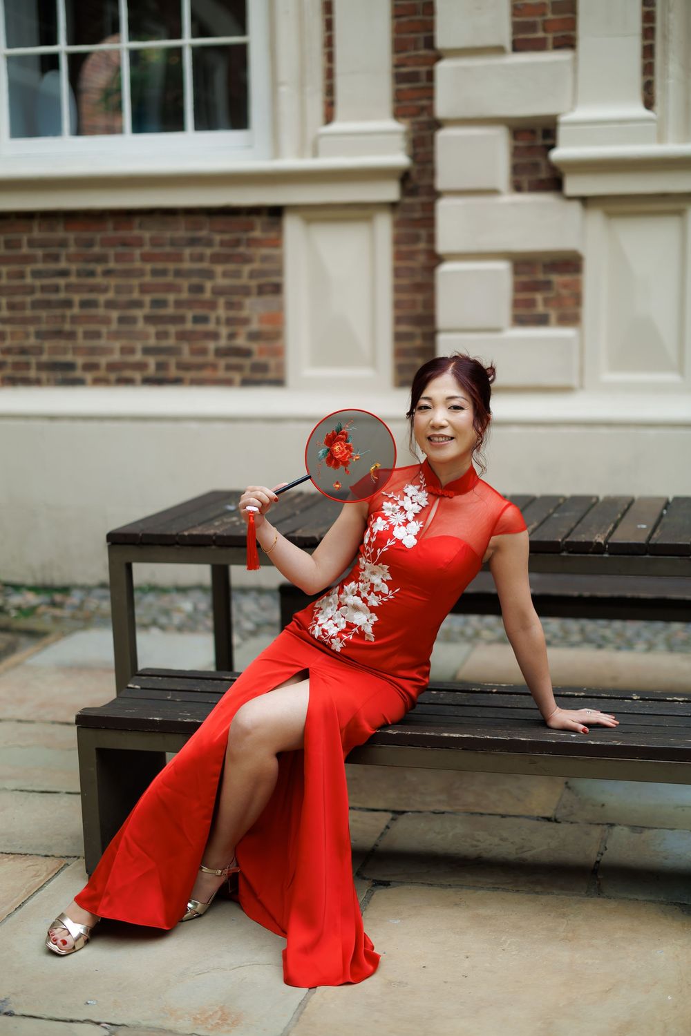 Woman in red cheongsam qipao dress holding decorative fan, seated on bench outside brick building.
