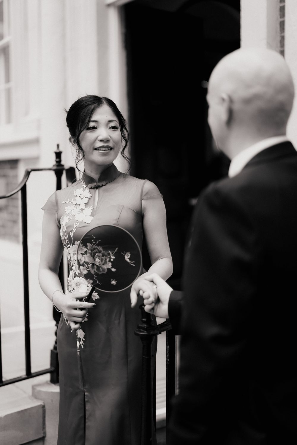 Elegant woman in traditional Chinese qipao dress holding a decorative fan, smiling at a man during a wedding ceremony.