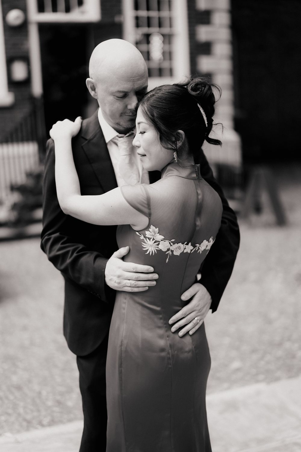 Black and white photo of a couple embracing, groom in suit holding bride in elegant backless floral dress.
