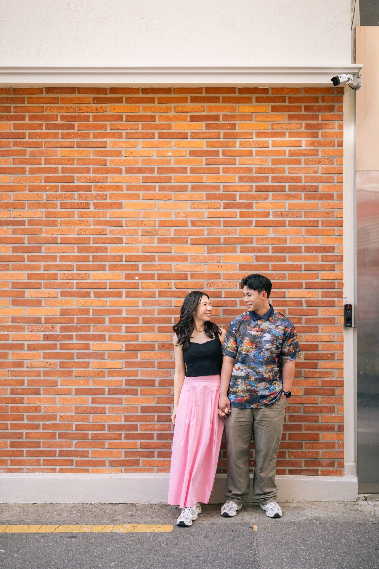 Young Asian couple holding hands and smiling at each other against a red brick wall outdoors.