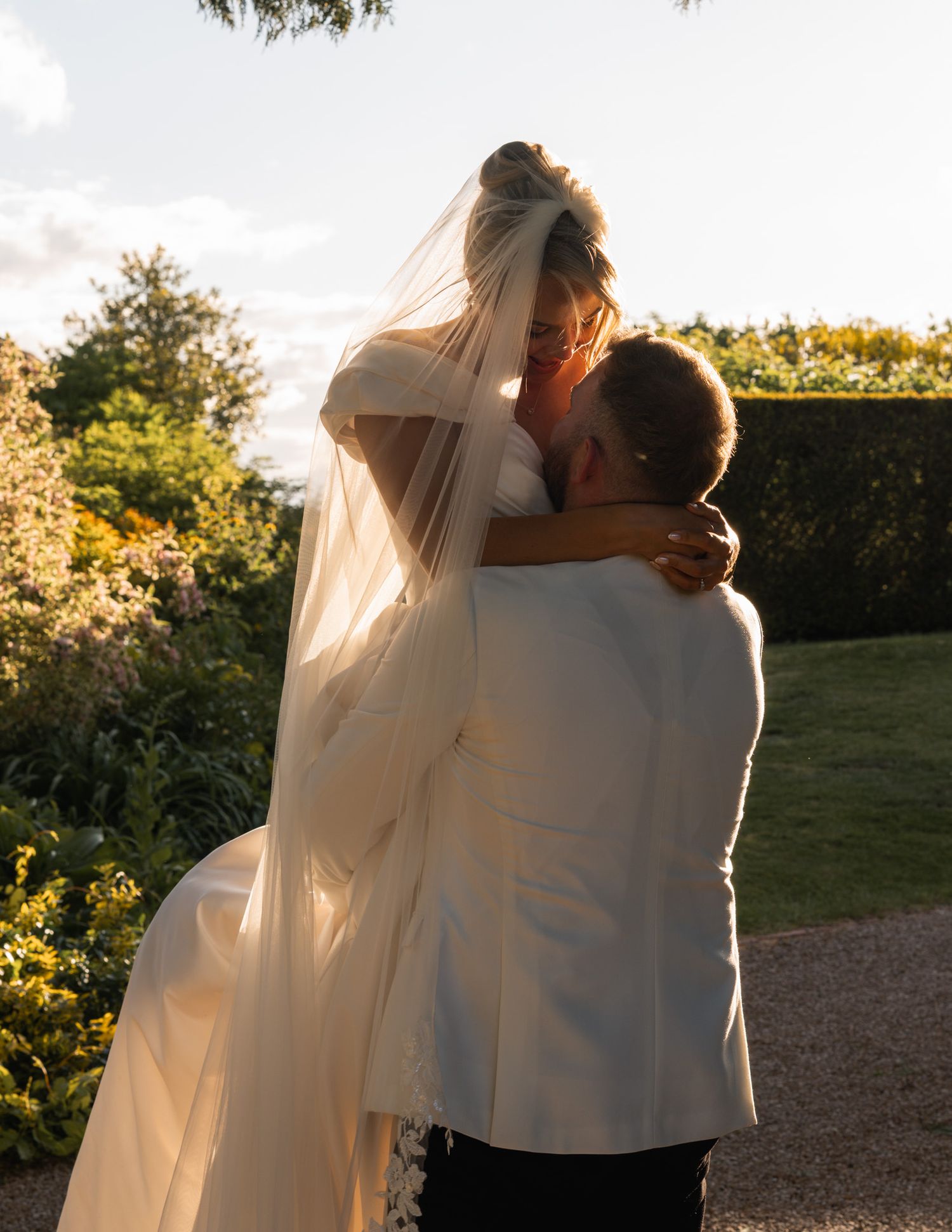 Bald groom in tweed suit and bride in satin gown smile while walking through a garden path.