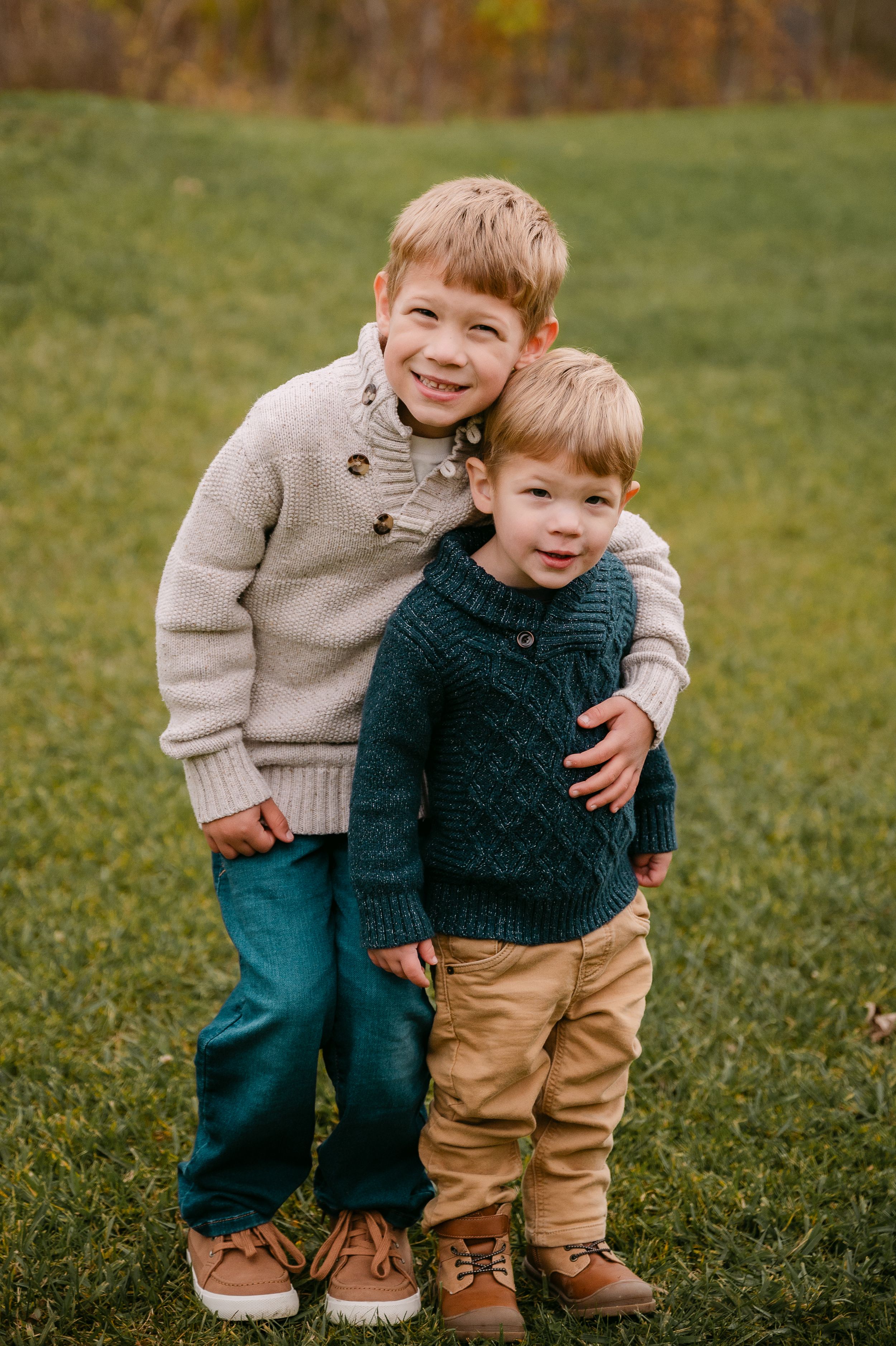 Two young brothers in knit sweaters share a warm hug while posing on a green grass field in autumn.