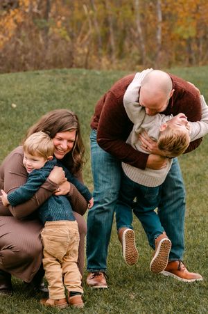 Happy family of four playing outdoors in autumn, parents hugging and lifting young children on green grass.