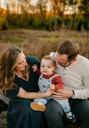 A young family with a baby boy sits outdoors in a golden autumn field, smiling warmly together.