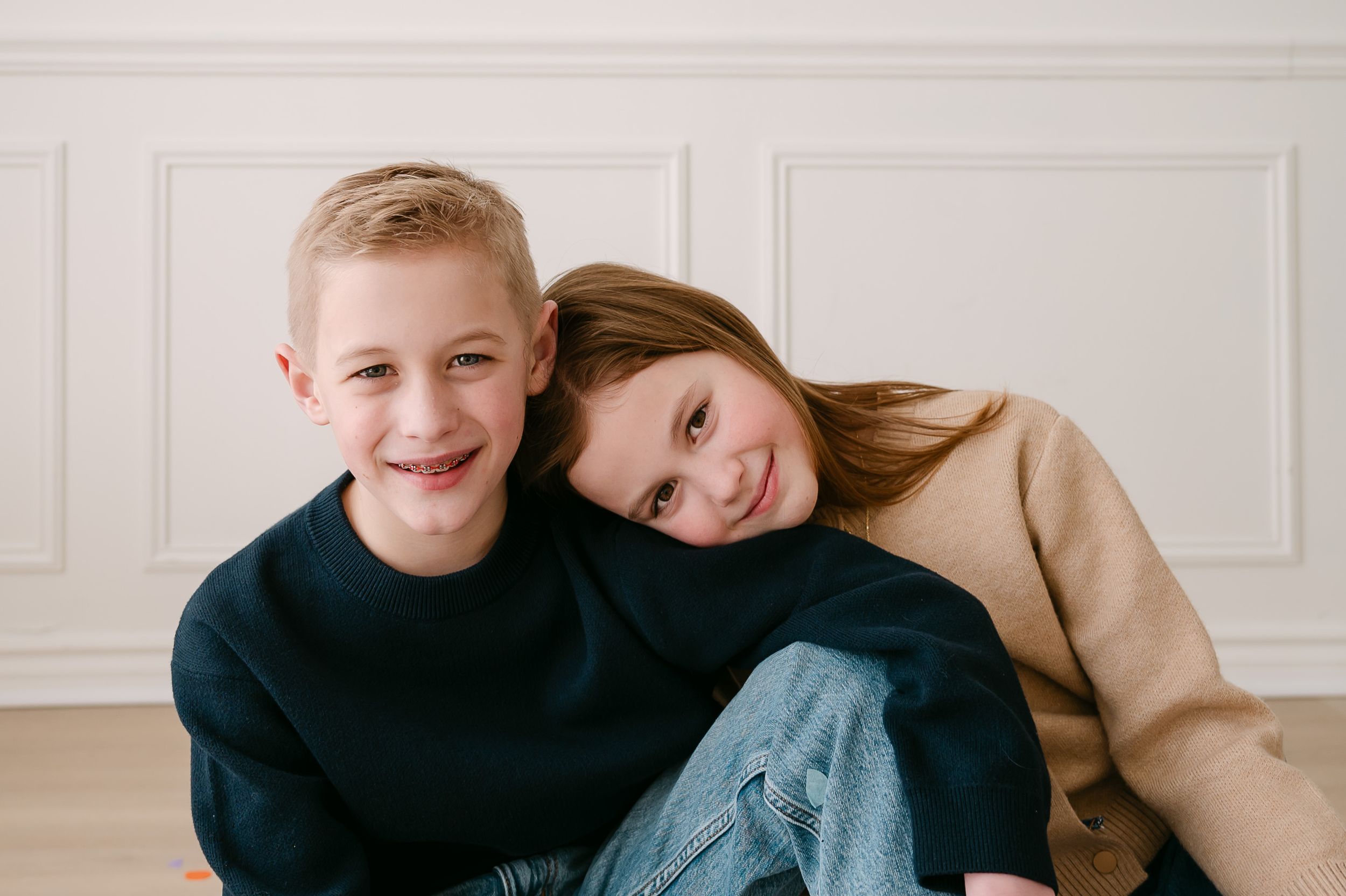 Two smiling children, a boy in navy and a girl in beige, pose together against a white wall.