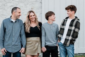 Happy family of four smiling and holding hands outdoors against a white wall in casual stylish outfits.