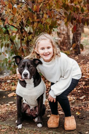 Young girl in white sweater smiling with black and white dog in matching sweater among autumn leaves.