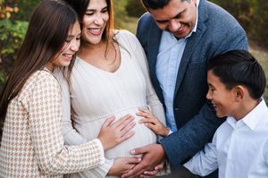 Family gathering around pregnant woman's belly, smiling outdoors in a warm maternity photo session.