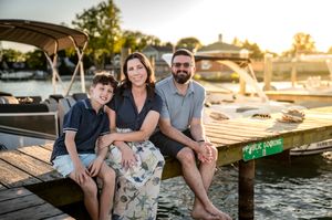 Family of three smiling together on a wooden dock at sunset with boats and water in the background.