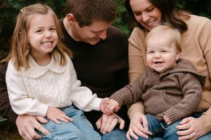 Happy family of four smiling outdoors, parents with young daughter and baby boy in cozy sweaters.