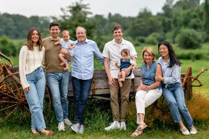 A multigenerational family smiling together outdoors near a rustic farm wagon surrounded by lush green fields.