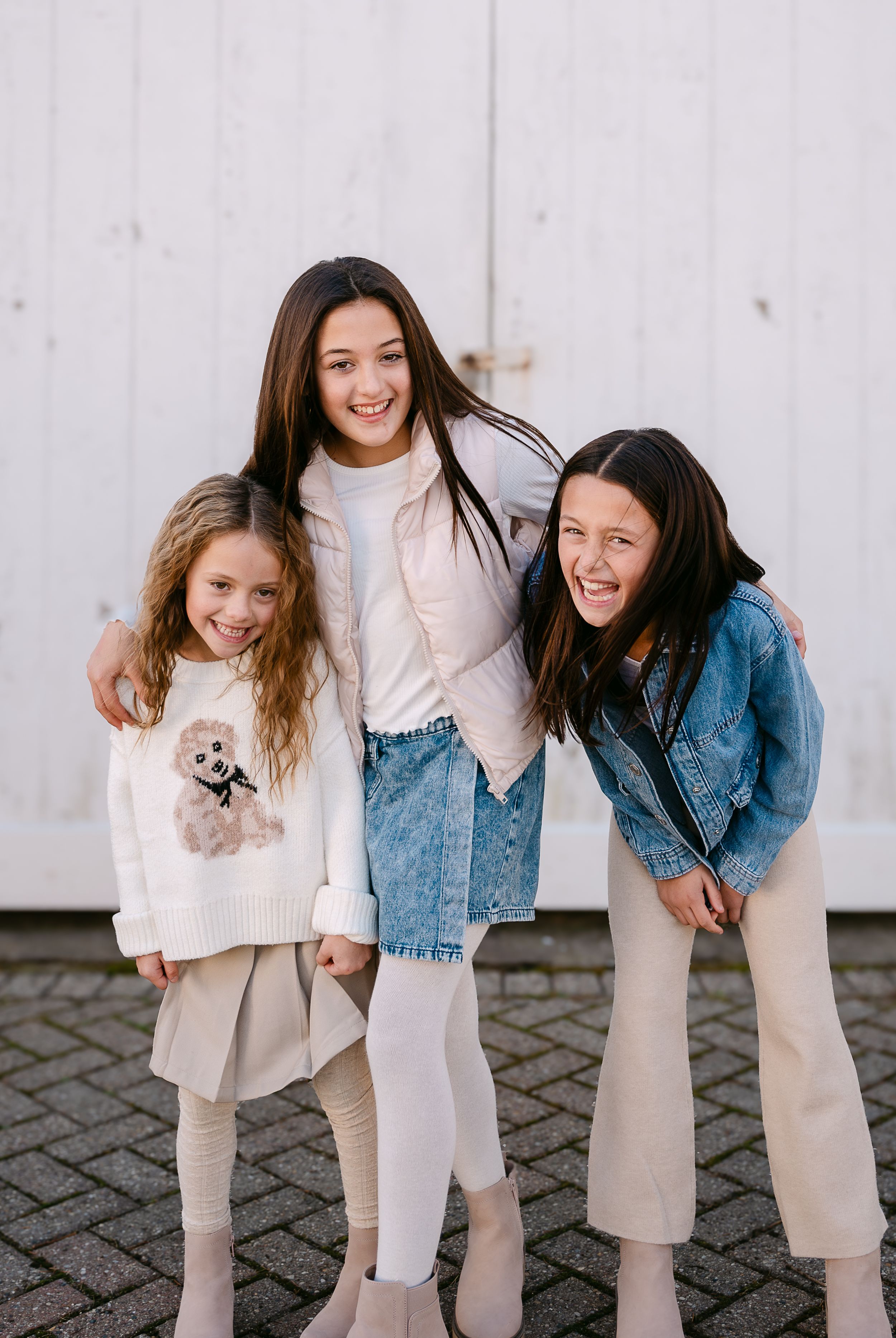 Three young girls smiling and posing together outdoors, wearing casual stylish outfits in neutral tones.