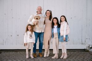 Family of four with a dog posing outdoors, smiling in coordinated neutral outfits against a white wall.