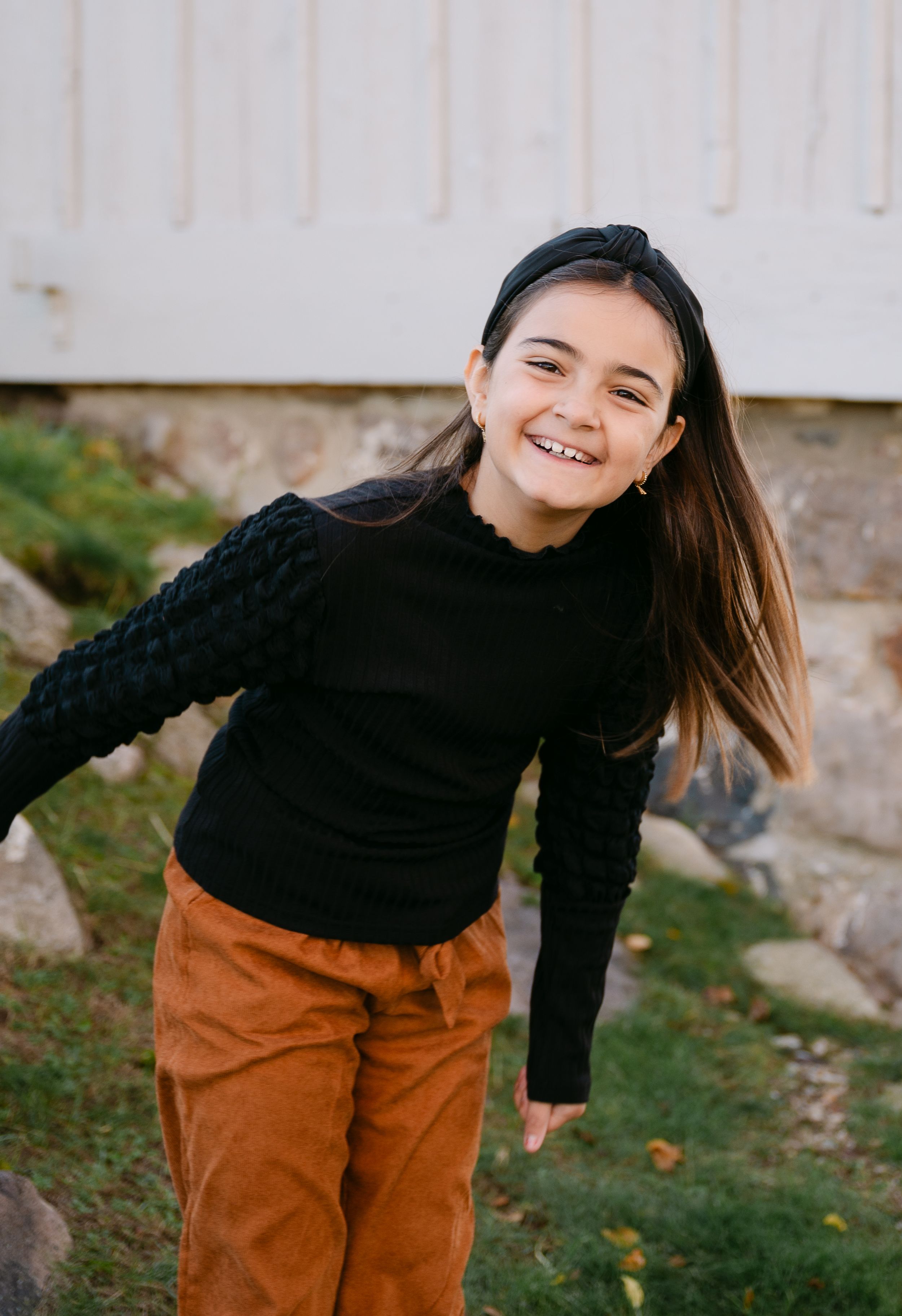 Smiling young girl in black sweater and brown pants running outdoors near stone wall.