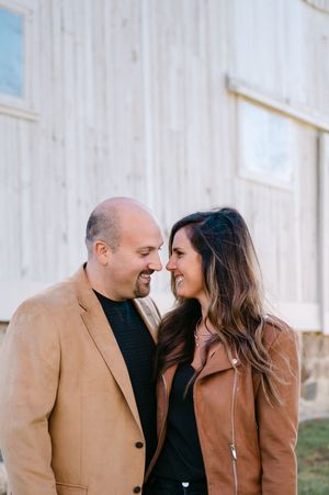 A smiling couple in tan jackets share a close romantic moment outdoors near a rustic white barn.