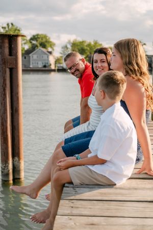 A family of four sits barefoot on a wooden dock, smiling by a calm waterway.