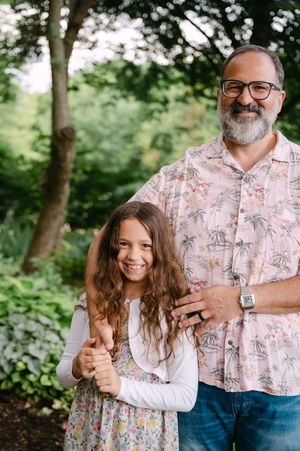 Smiling bearded father with glasses posing with young curly-haired daughter in a lush garden.