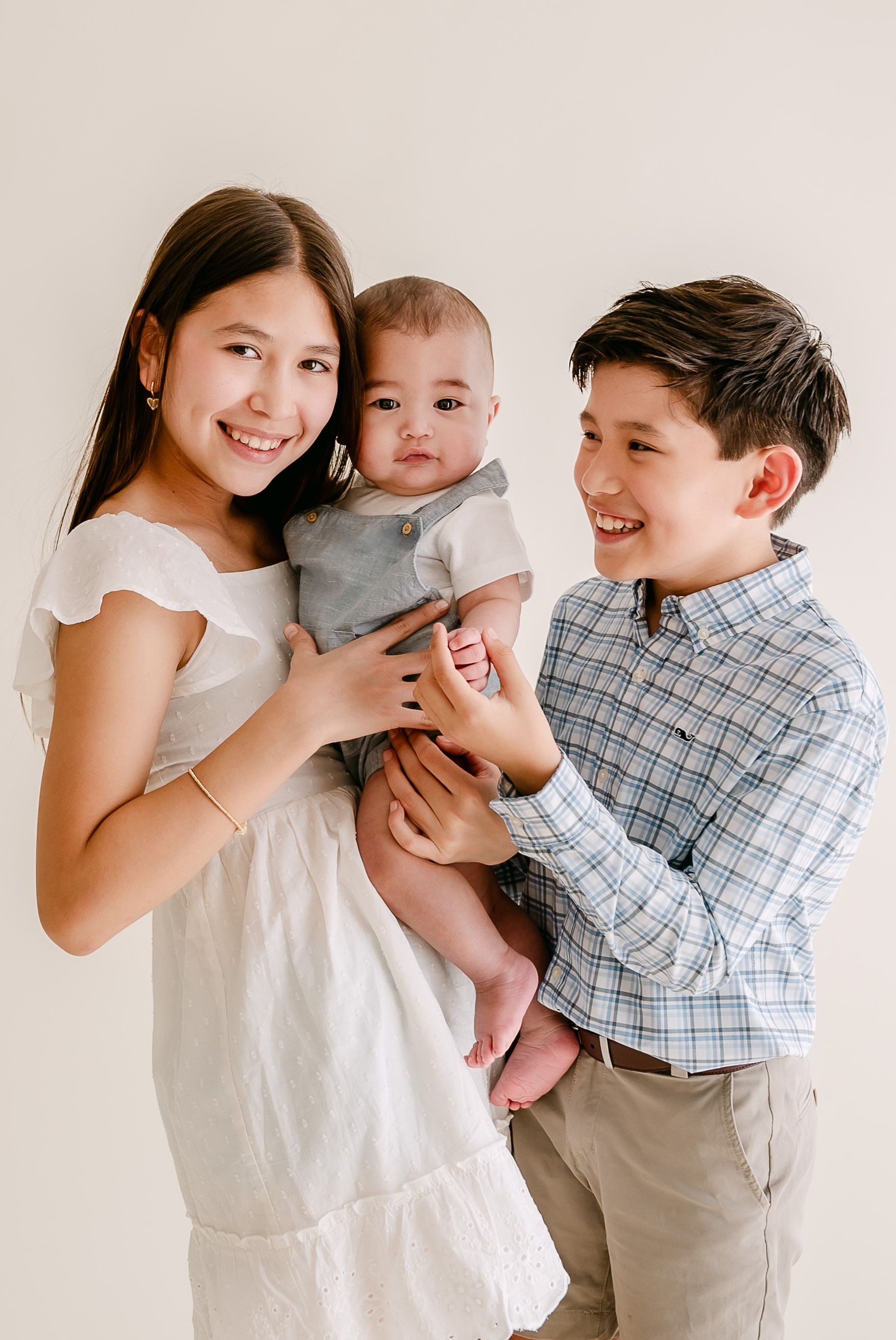 Two siblings smiling and holding a baby in a bright studio portrait, dressed in white, plaid, and gray outfits.