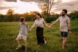 Family of three holding hands walking through green field at golden sunset hour.