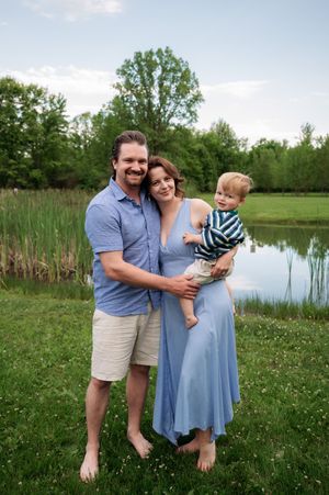 A smiling family of three poses barefoot by a pond, with a man in blue, a pregnant woman in a blue dress holding a toddler.