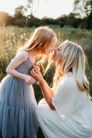Blonde mother and red-haired daughter sharing a tender nose-to-nose moment in a sunlit meadow.