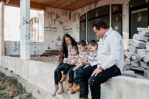 Family of five sitting on construction site ledge, parents smiling with three young children in matching outfits.