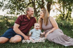 A couple sits in green grass smiling at their happy baby during a golden sunset session.