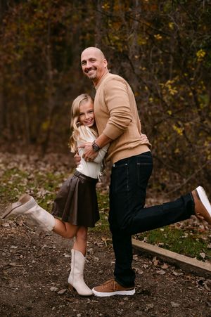 A bald man and young girl hug and smile outdoors in fall, she wears white boots and brown skirt.