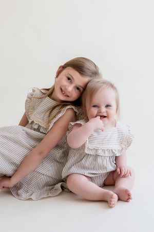 Two young sisters in matching striped ruffle dresses pose together, smiling, against a white background.