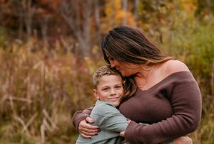 A mother hugs her smiling young son outdoors during autumn, surrounded by colorful fall foliage.