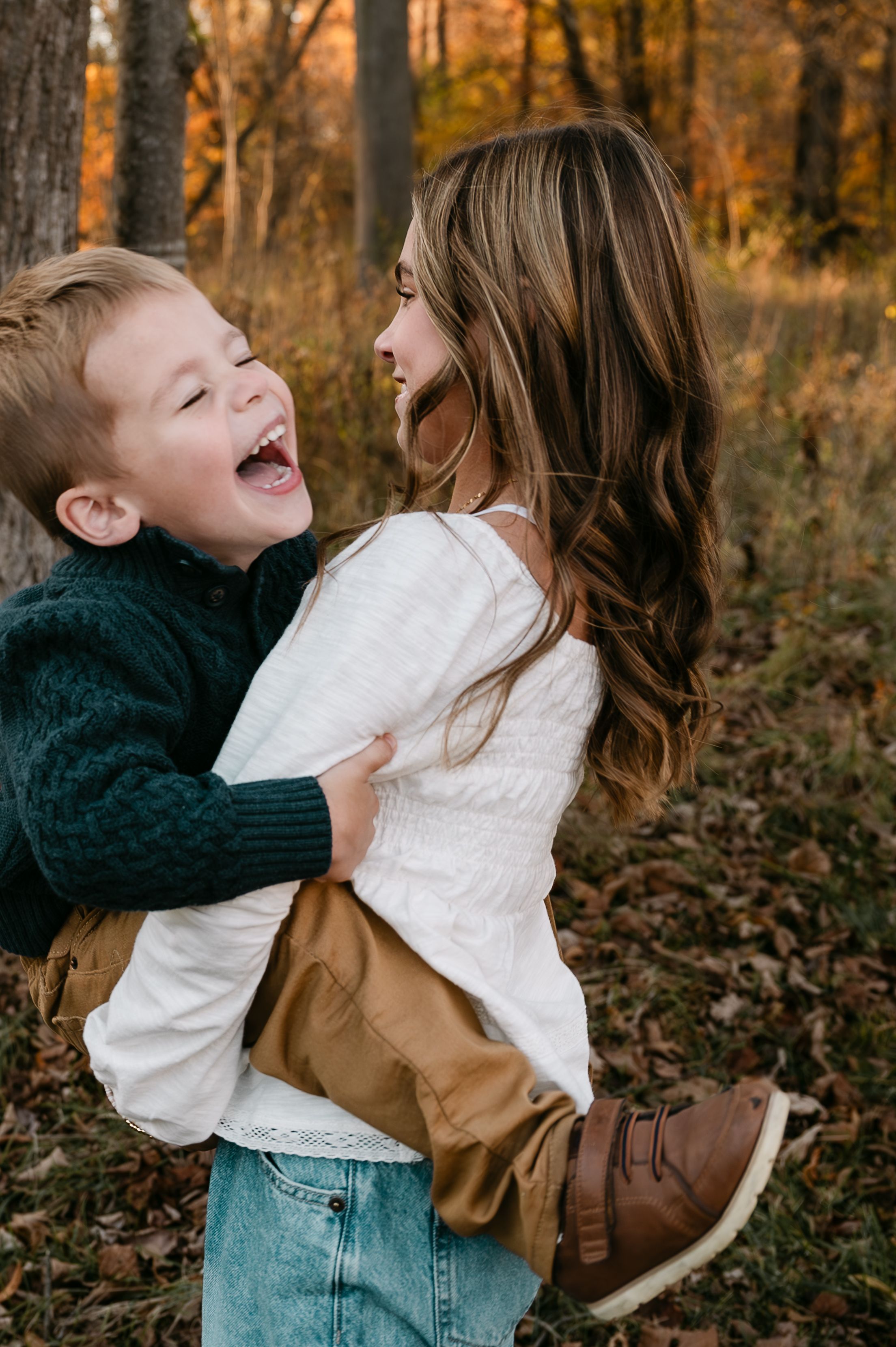 A laughing boy and girl share a joyful hug outdoors in an autumn forest with colorful fall foliage.