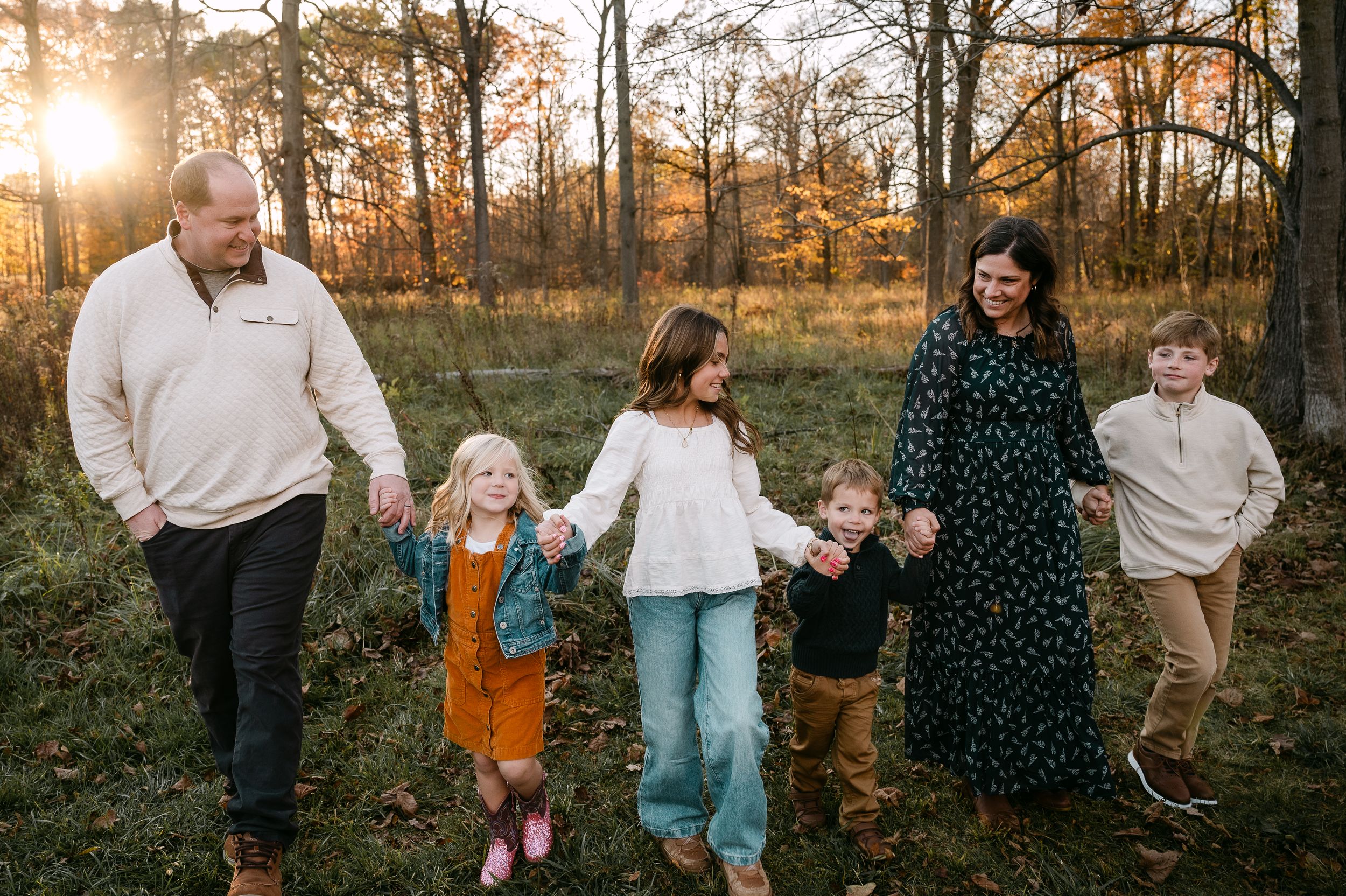 Family of six walking hand-in-hand through a sunlit autumn field at golden hour.