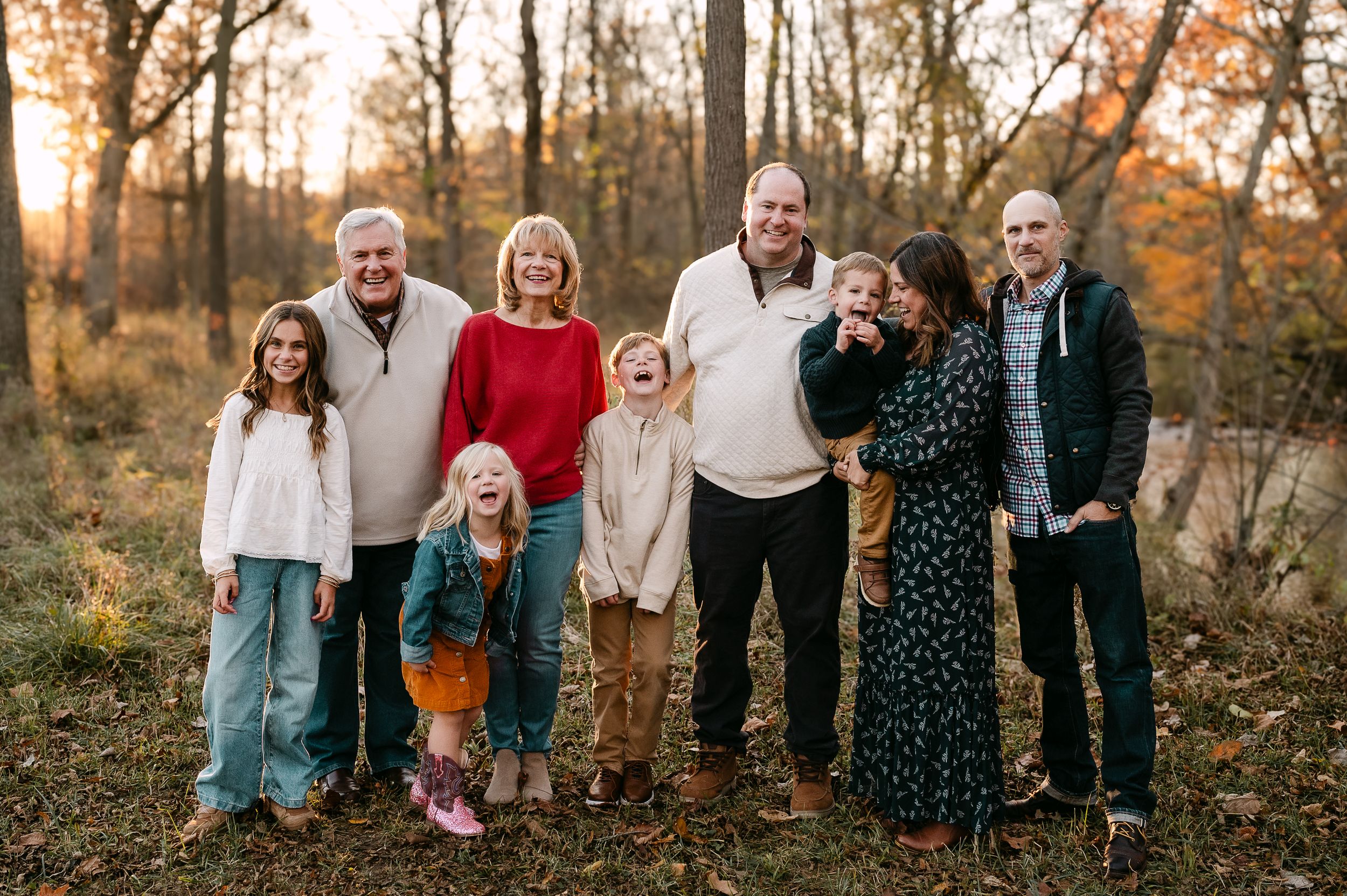 Multi-generational family of eight smiling together outdoors in an autumn forest setting during golden hour.