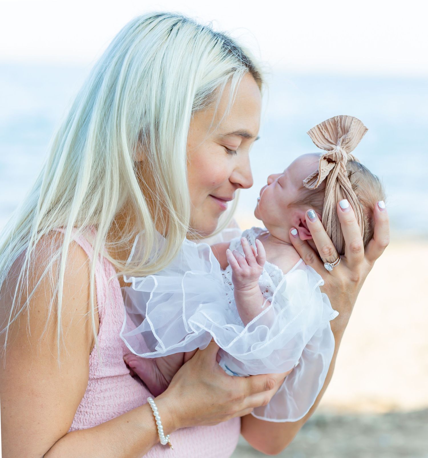 Blonde woman tenderly holding a newborn baby in a white ruffle outfit with bow headband at the beach.