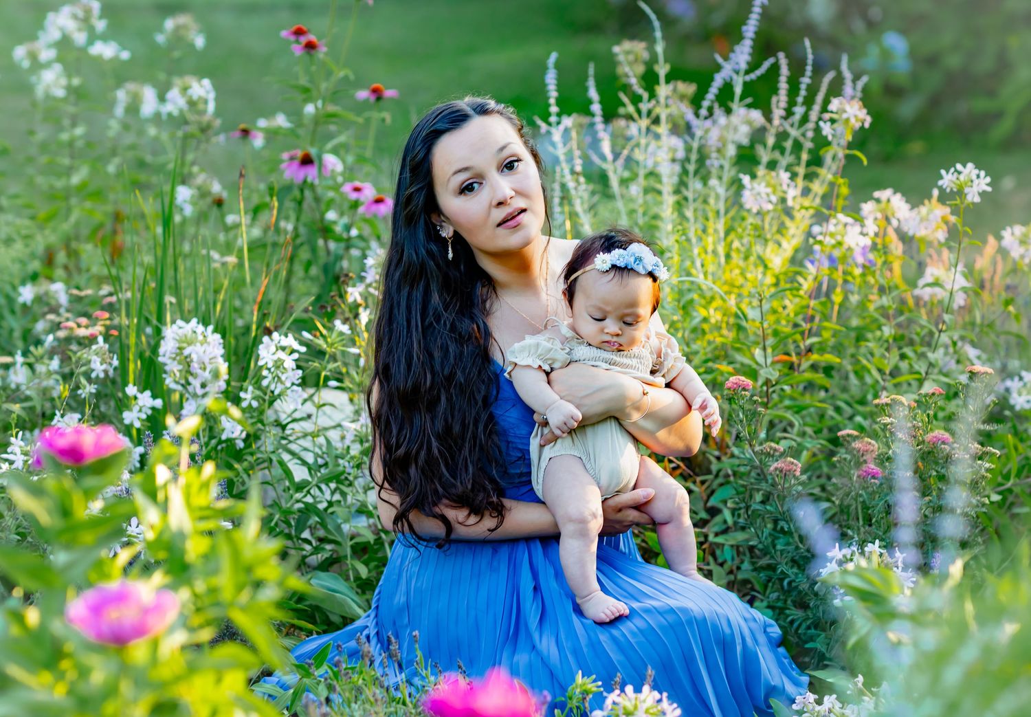 Woman in blue dress holding baby girl among colorful wildflowers in a lush garden setting.