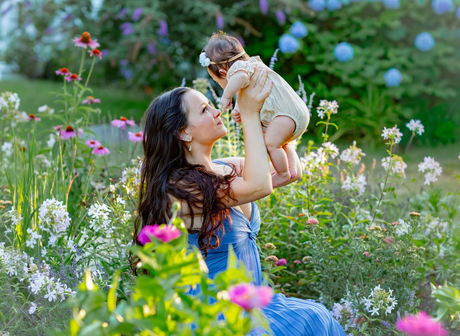 A mother joyfully lifts her baby in a lush garden filled with colorful blooming flowers on a sunny day.