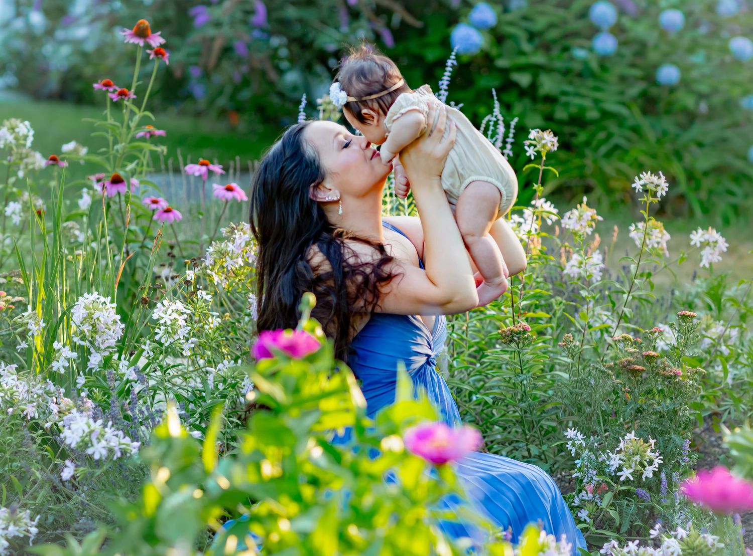 A mother lovingly lifts her baby among colorful wildflowers in a lush garden setting.