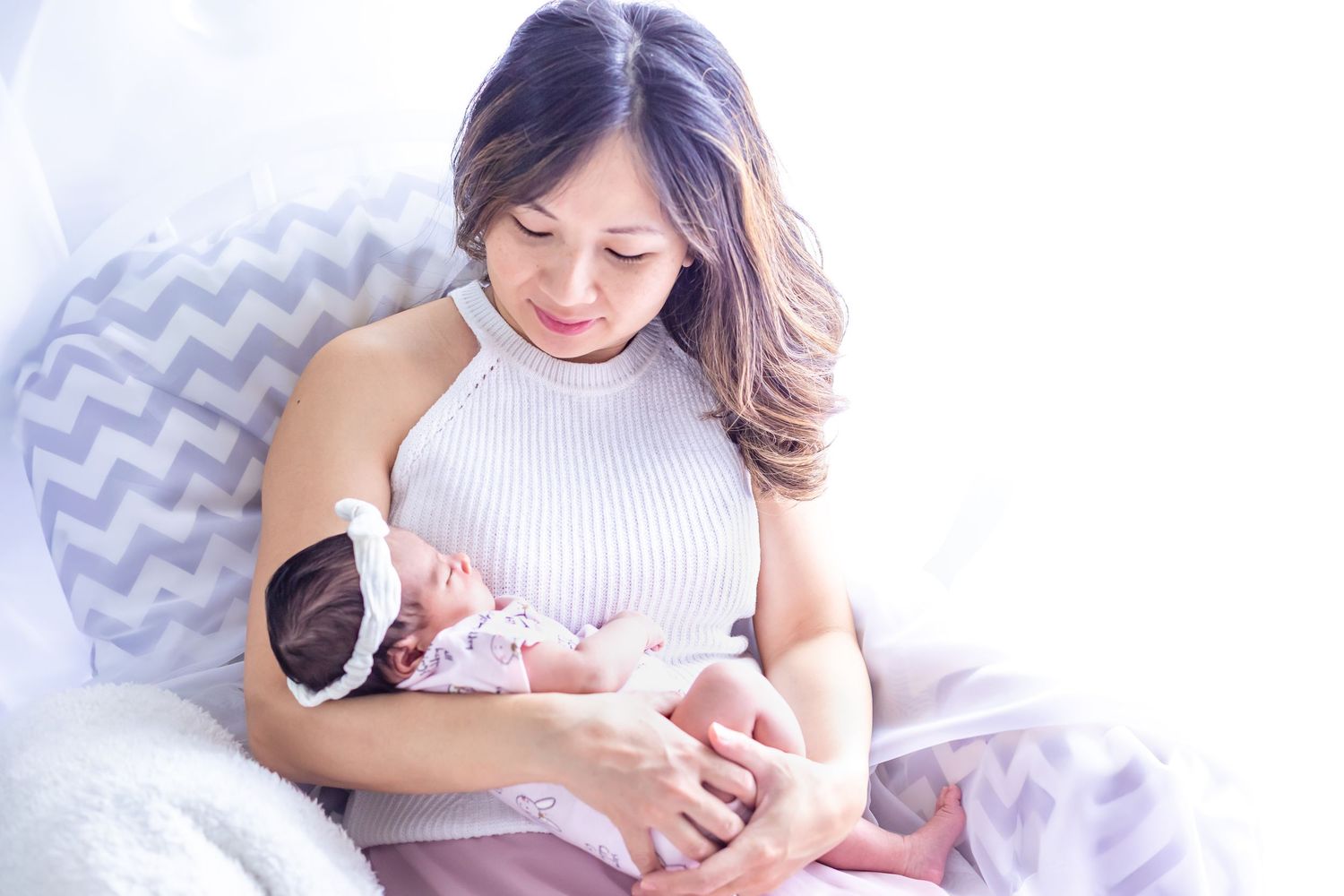 A smiling Asian mother tenderly holds her sleeping newborn baby while seated in a cozy chair.