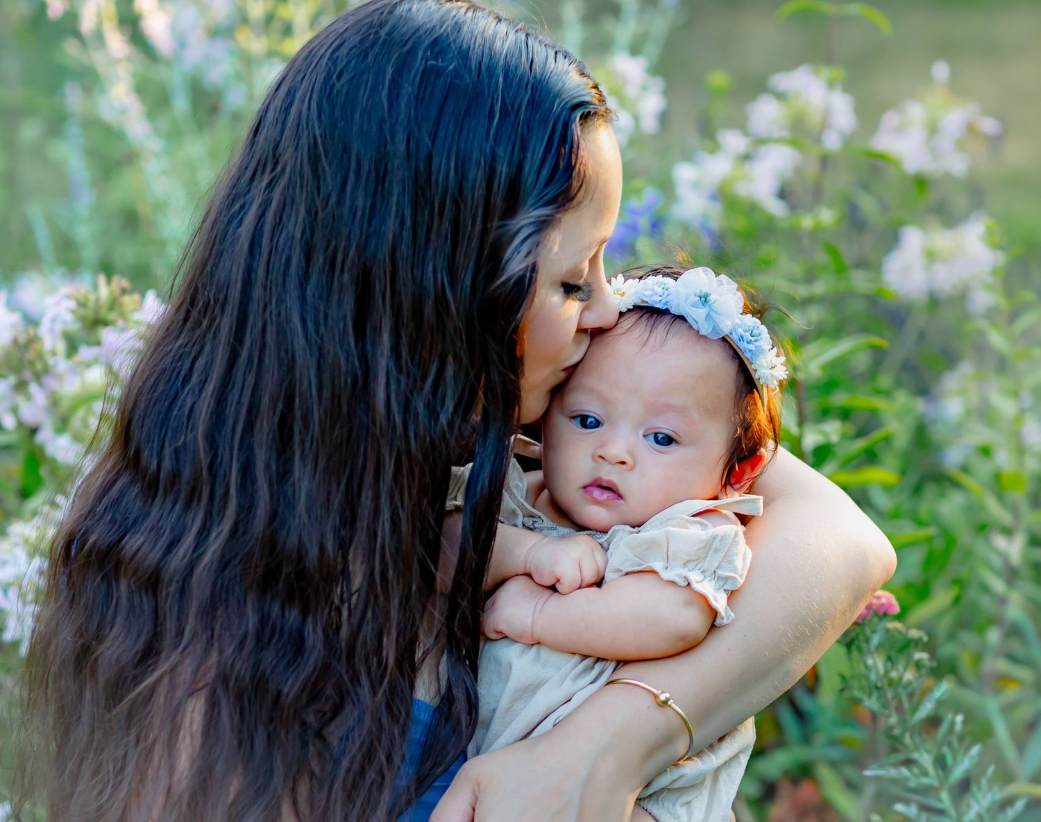 Mother kissing baby girl wearing floral headband outdoors among soft purple wildflowers.