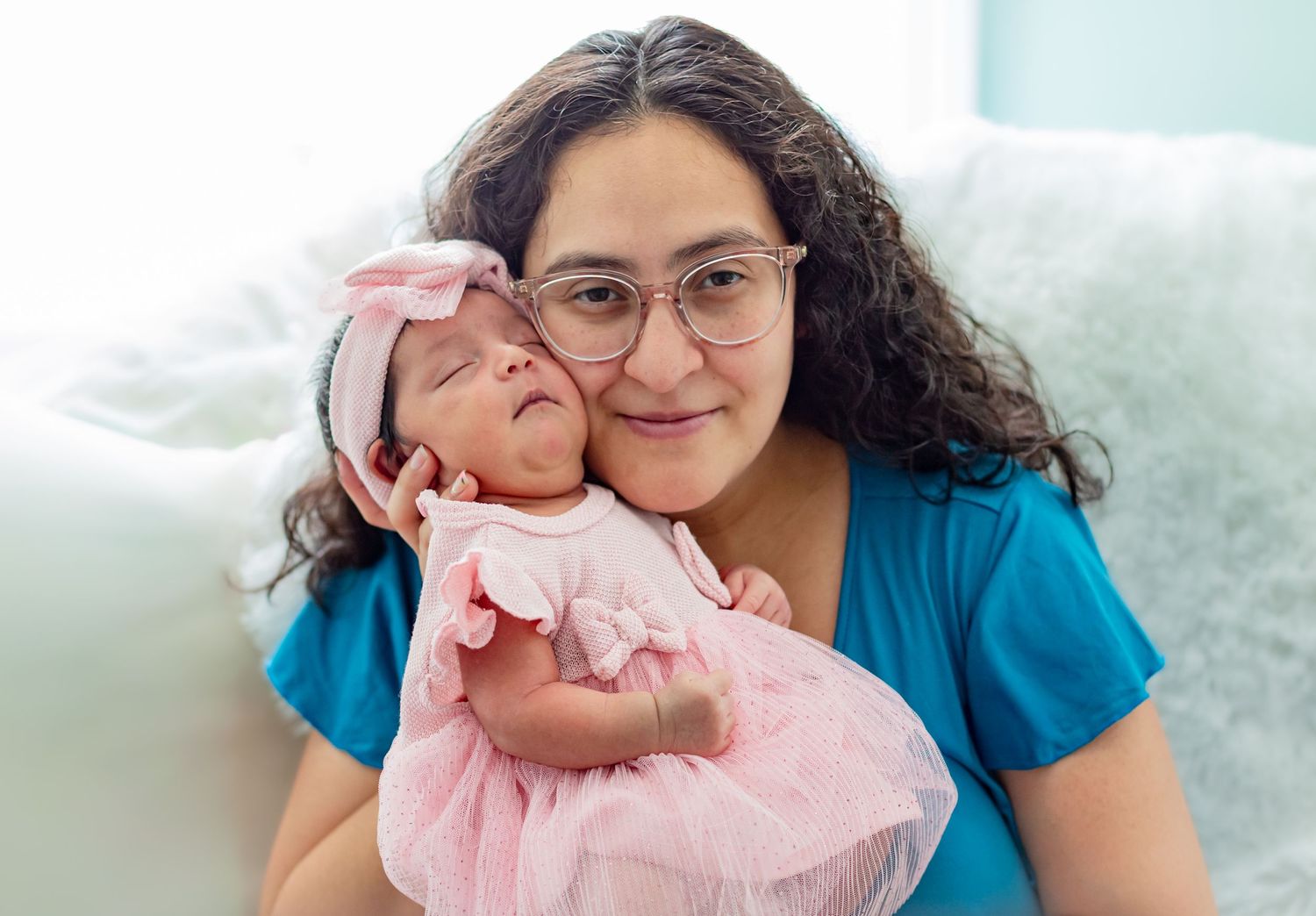 A smiling woman with glasses holds a newborn baby in a pink dress and headband, posing for a portrait.