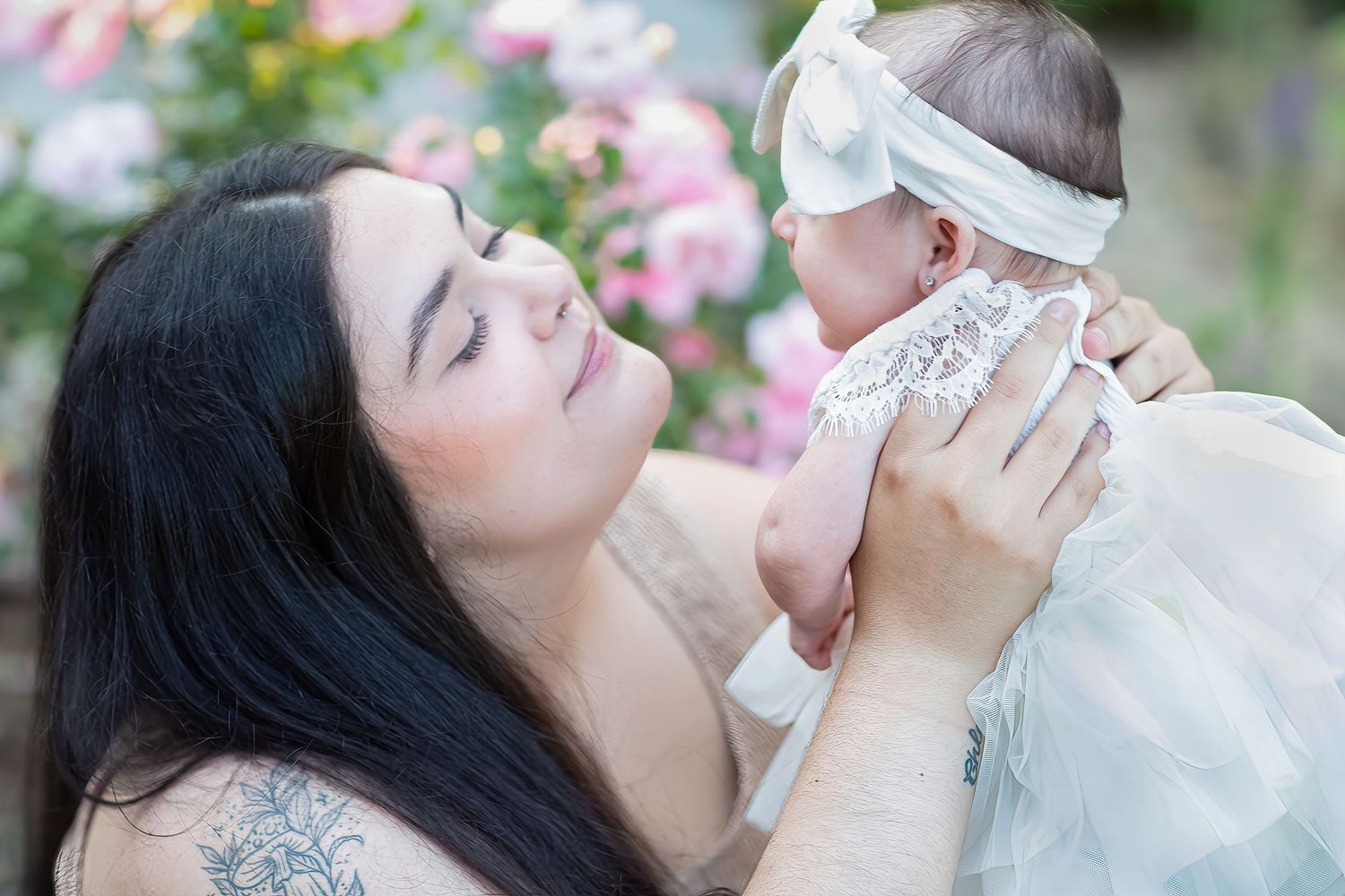 A mother with dark hair and tattoos lovingly holds her newborn baby in a white dress outdoors near pink flowers.