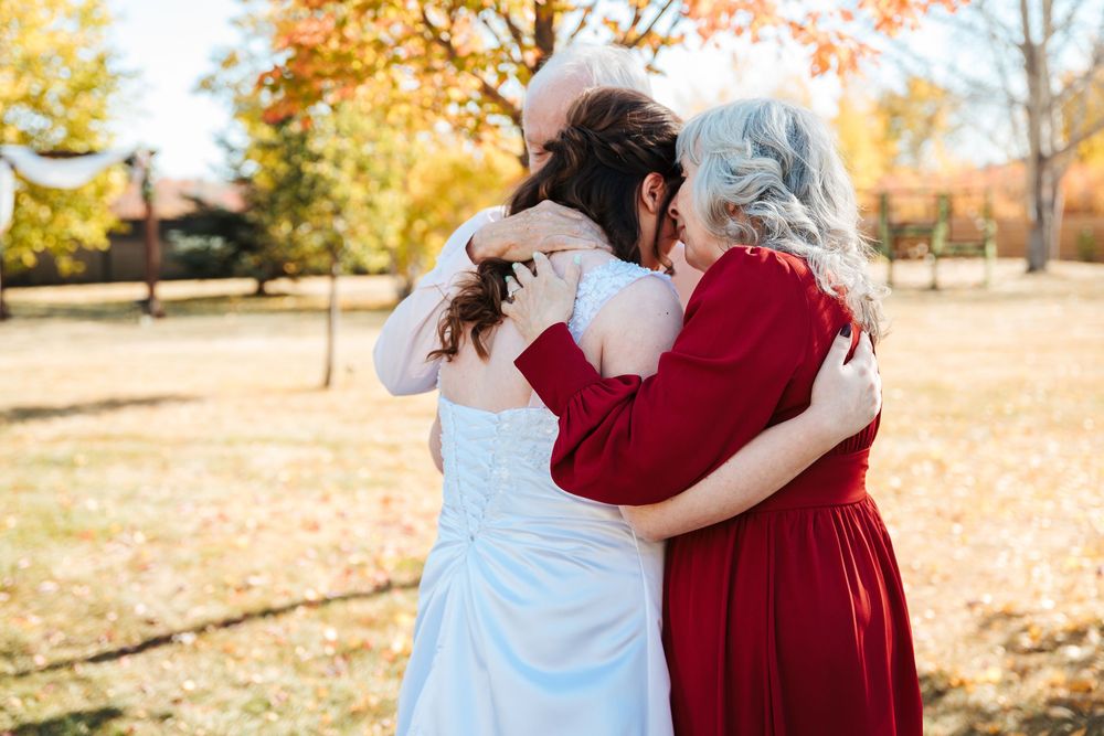Bride in white gown embracing two women in red dresses outdoors amid autumn foliage fall wedding celebration.