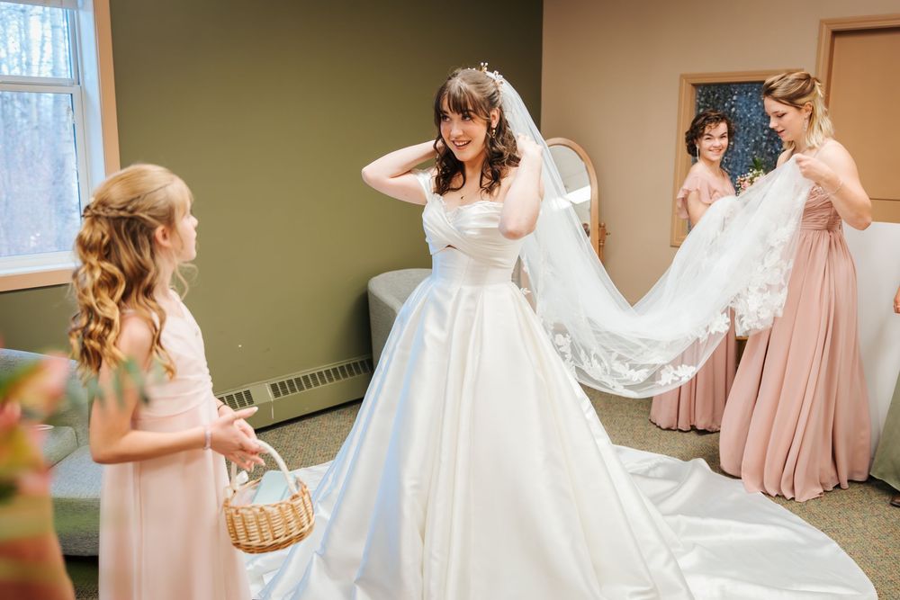 Bride in white gown adjusts veil while bridesmaids in pink dresses and flower girl assist in bridal suite.
