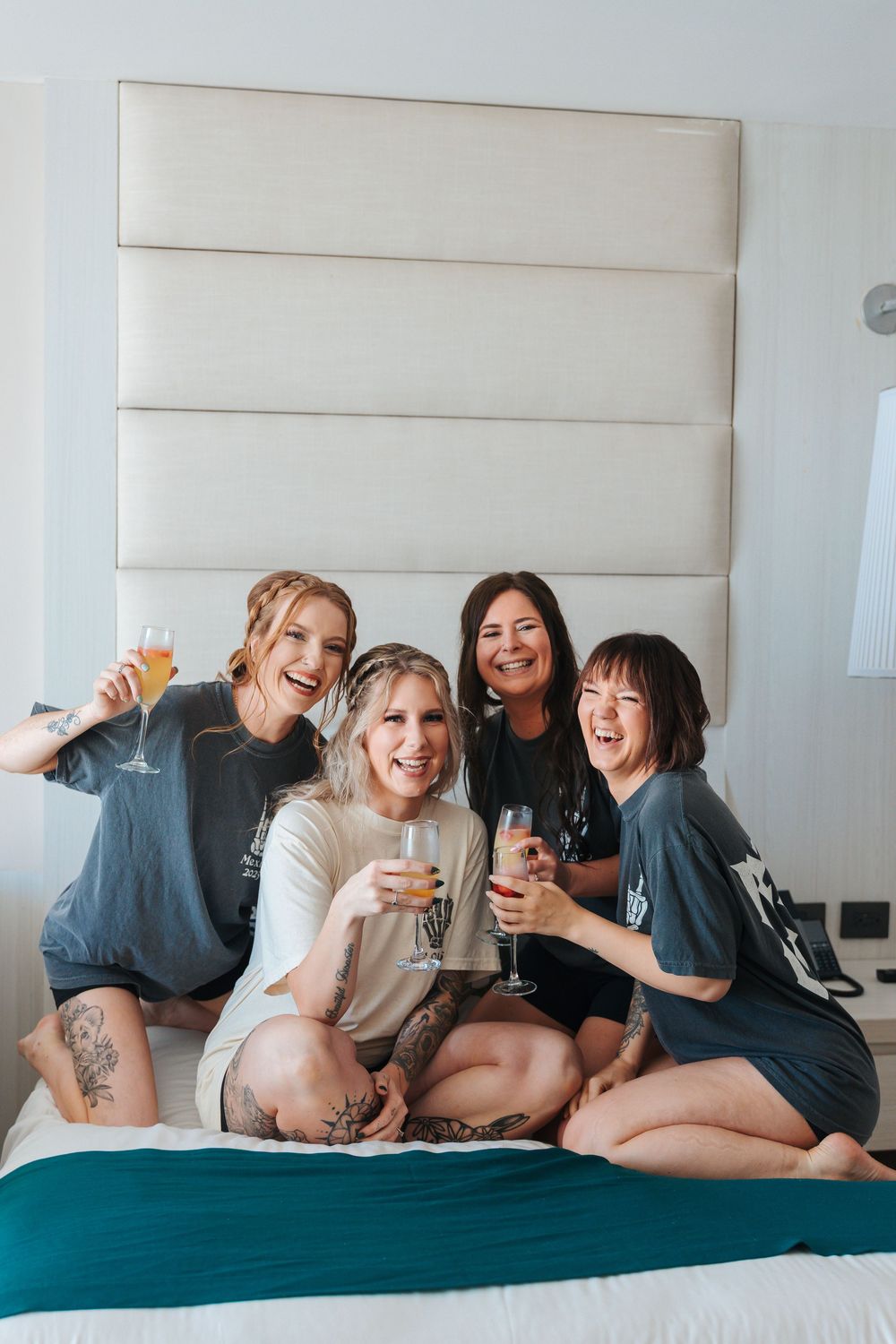 Four bridesmaidslaughing and holding champagne glasses while sitting on a hotel bed in matching gray shirts. mexico wedding resorts